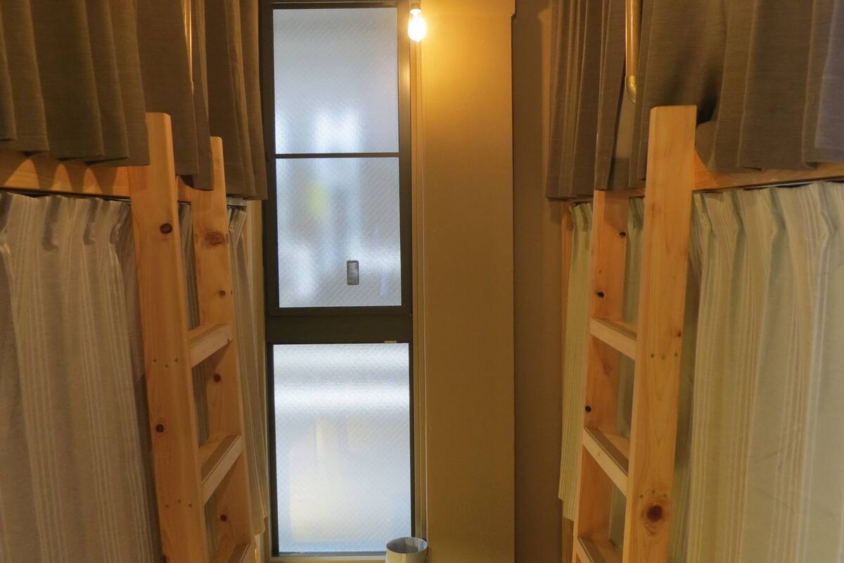 A narrow hallway with wooden bunk beds on either side, a window at the end, and a light bulb hanging from the ceiling.