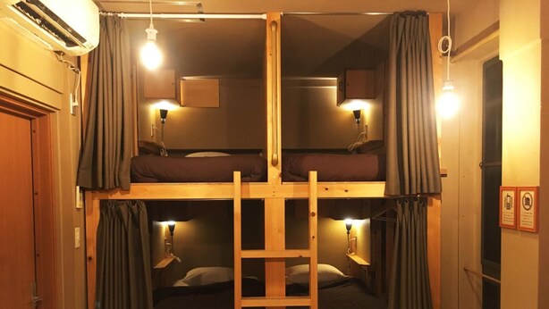 A bunk bed room with two beds, a ladder, bedside lamps, and an air conditioning unit. - No Borders Hostel (Tokyo)