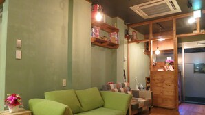 A cozy living room with a green sofa, a small table with a vase of flowers, and wooden shelves with books and decorations. - No Borders Hostel (Tokyo)