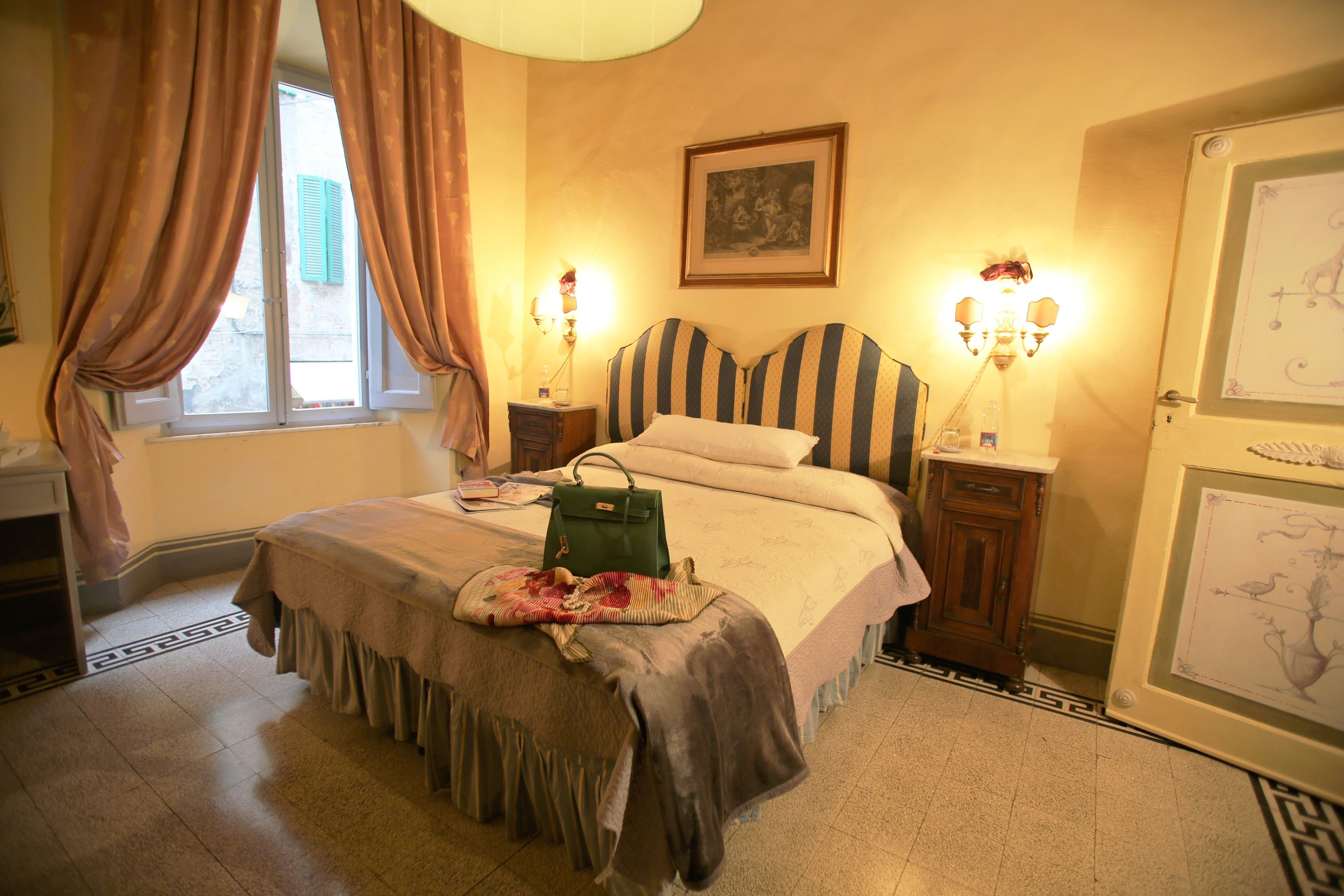 A bedroom with a bed, a bedside table, a lamp, a window with curtains, and a wall-mounted picture.