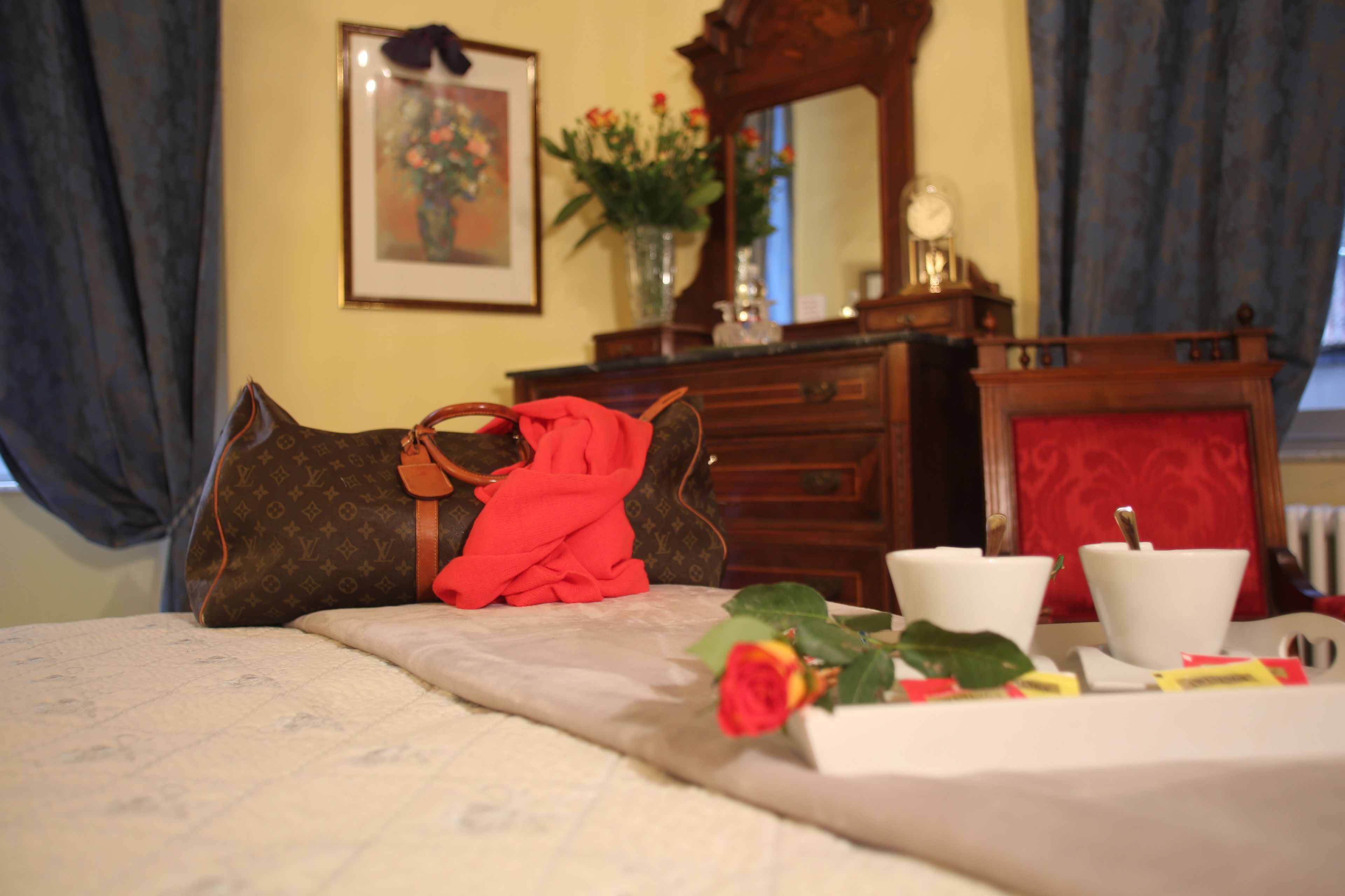 Un lit bien fait, recouvert d’un tissu rouge, un sac Louis Vuitton marron, un vase rempli de fleurs et un miroir posé sur une commode.