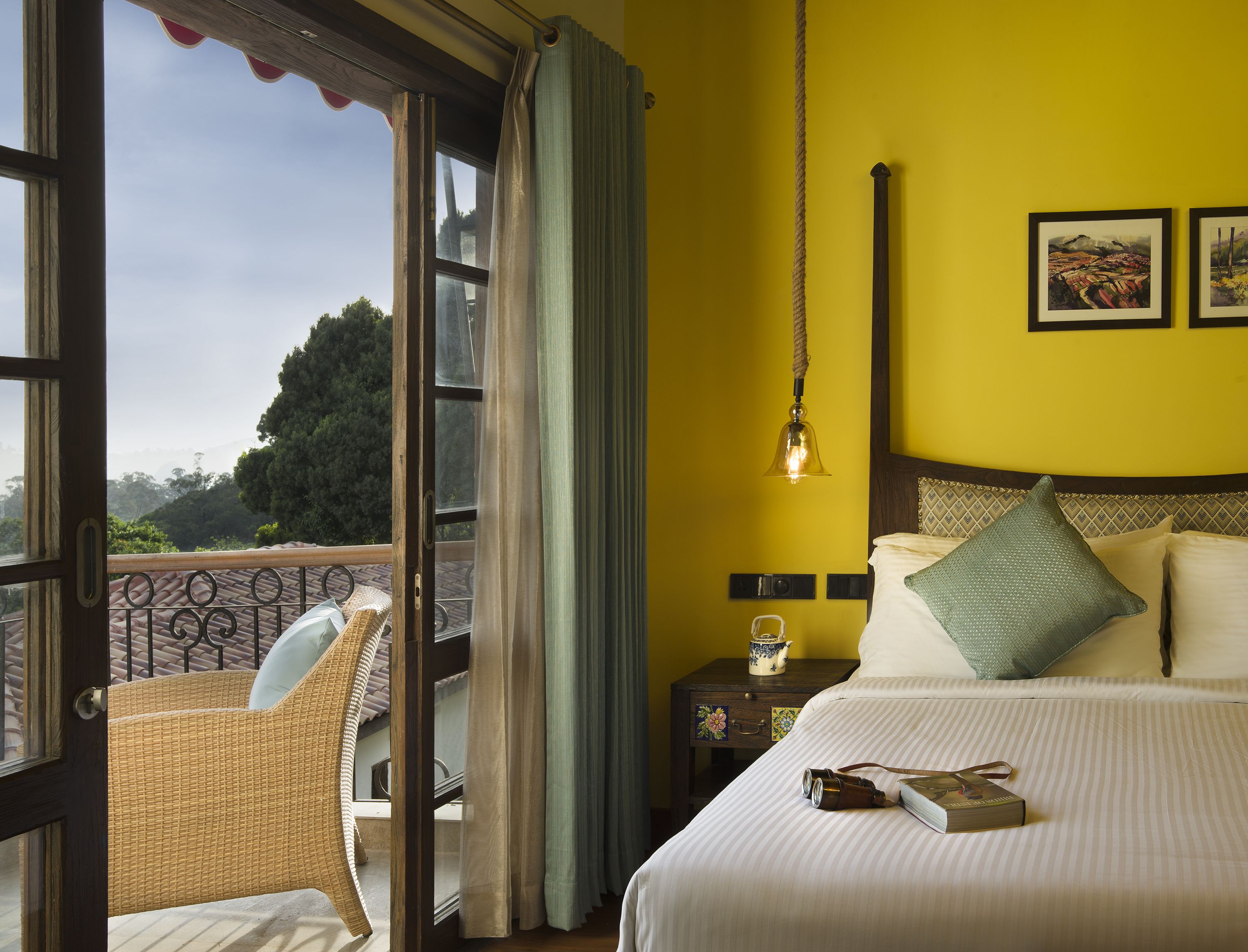 A room with a bed, a nightstand, a wicker chair, a window with curtains, and a view of the outdoors.