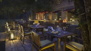Un espacio de comedor al aire libre con mesas y sillas, un edificio de piedra al fondo y vista al agua.