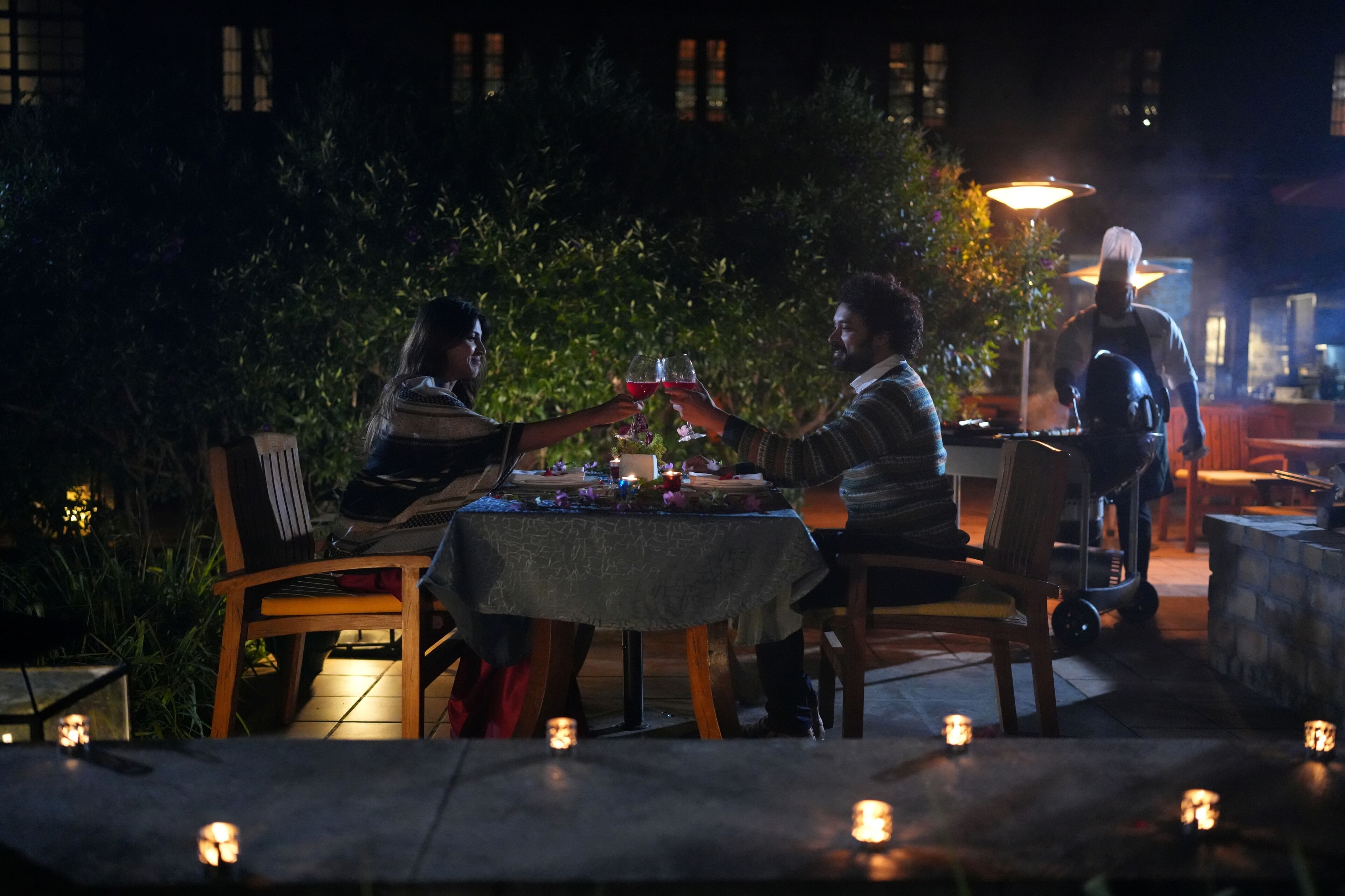 An outdoor dining area with a table set for two, surrounded by candles, and a waiter in the background.