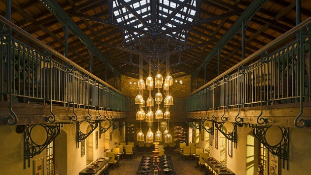 Un restaurante con techo alto, barandillas de metal ornamentadas y candelabros de cristal colgantes. Las mesas están dispuestas en el centro con sillas a cada lado.
