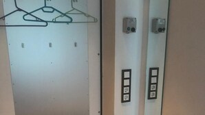 Una habitación con una silla blanca, un espejo, un interruptor de luz en la pared y una puerta con un panel de vidrio satinado y perchas.
