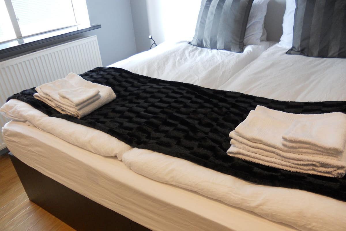 A neatly made bed with a black blanket and white pillows, a radiator, and a window with blinds.