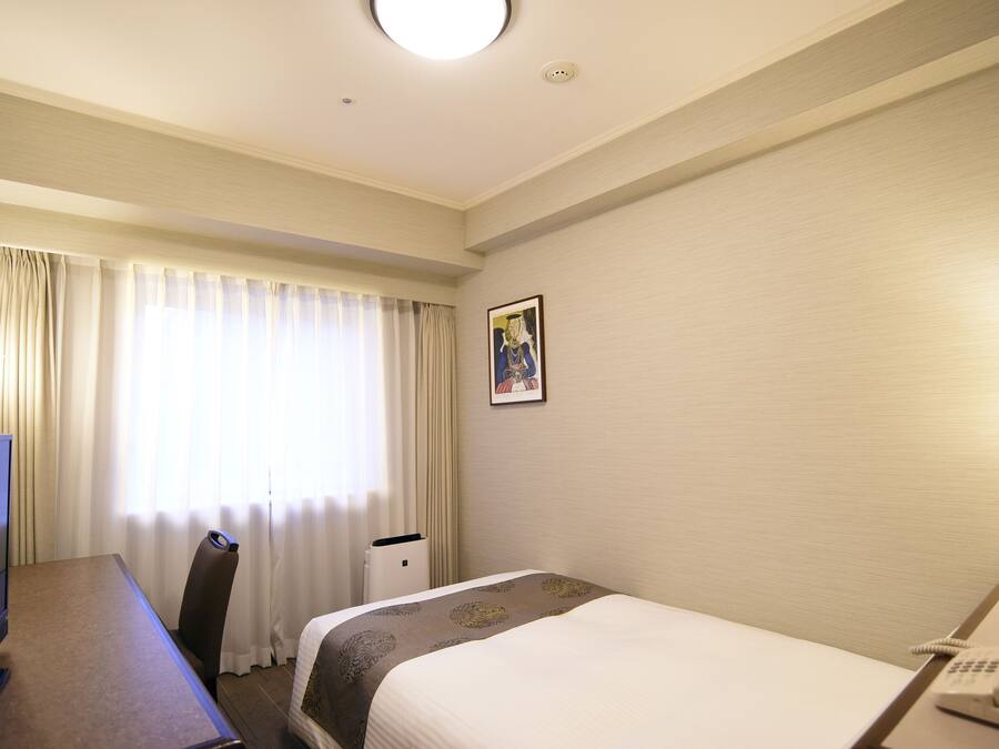 Una camera d'albergo con un letto, una scrivania, una sedia, una lampada, un telefono e una finestra con tende.