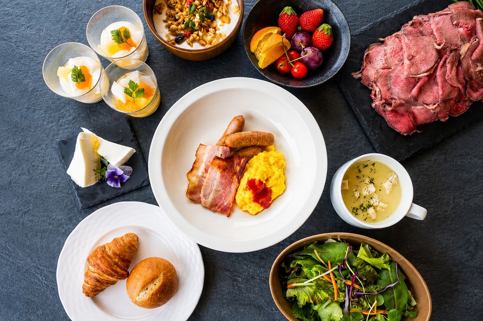 A breakfast setting with plates of food, including eggs, bacon, and toast, alongside a bowl of salad and a cup of soup.