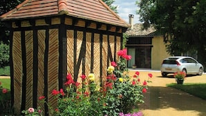 A traditional wooden gazebo with a tiled roof, surrounded by vibrant flower beds and a pathway leading to a parked car. - Les Sureaux (Le Pizou)