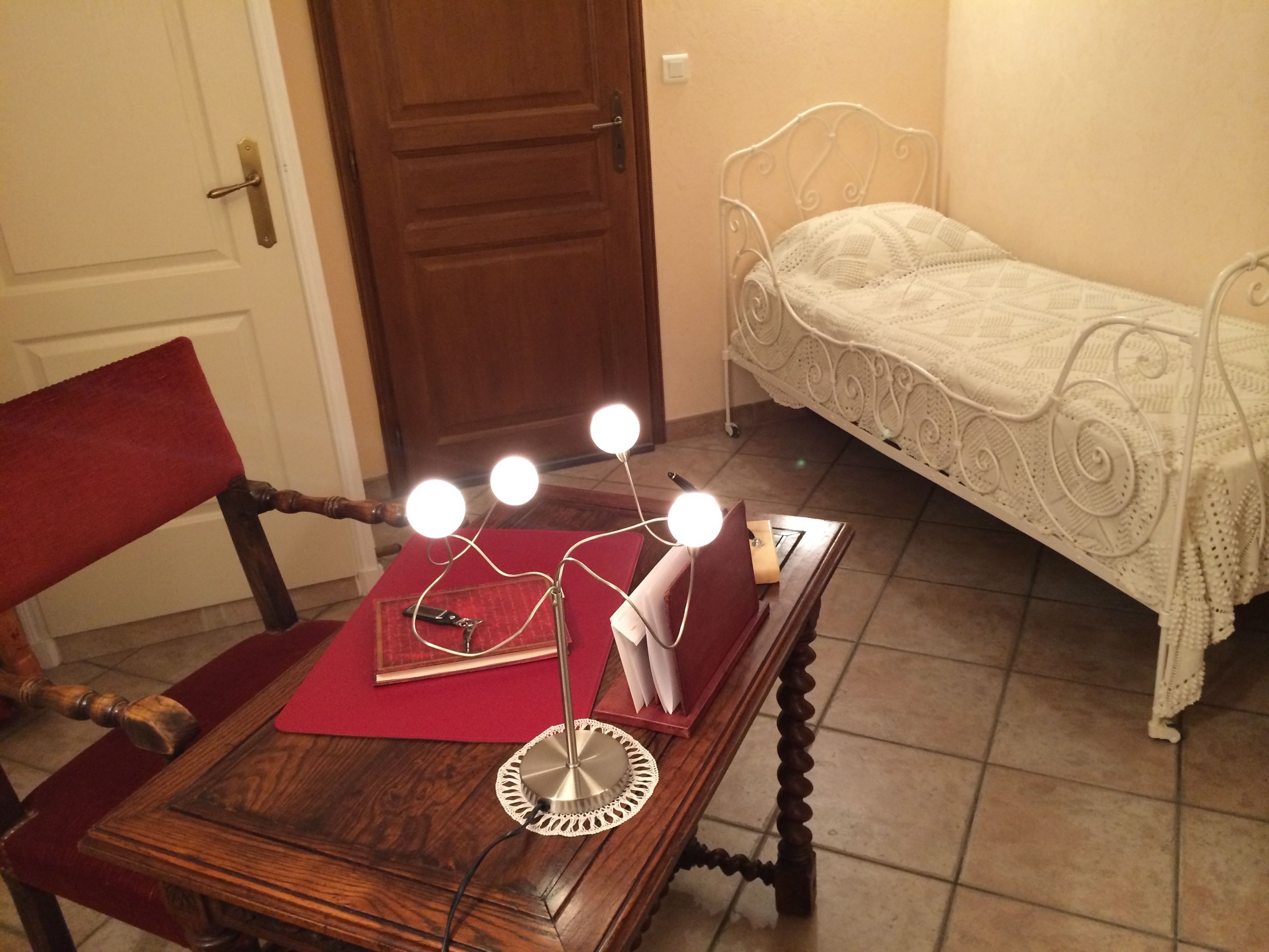 A small bedroom with a wooden table, a red chair, a white metal bed frame, and a bedside table with three lights.