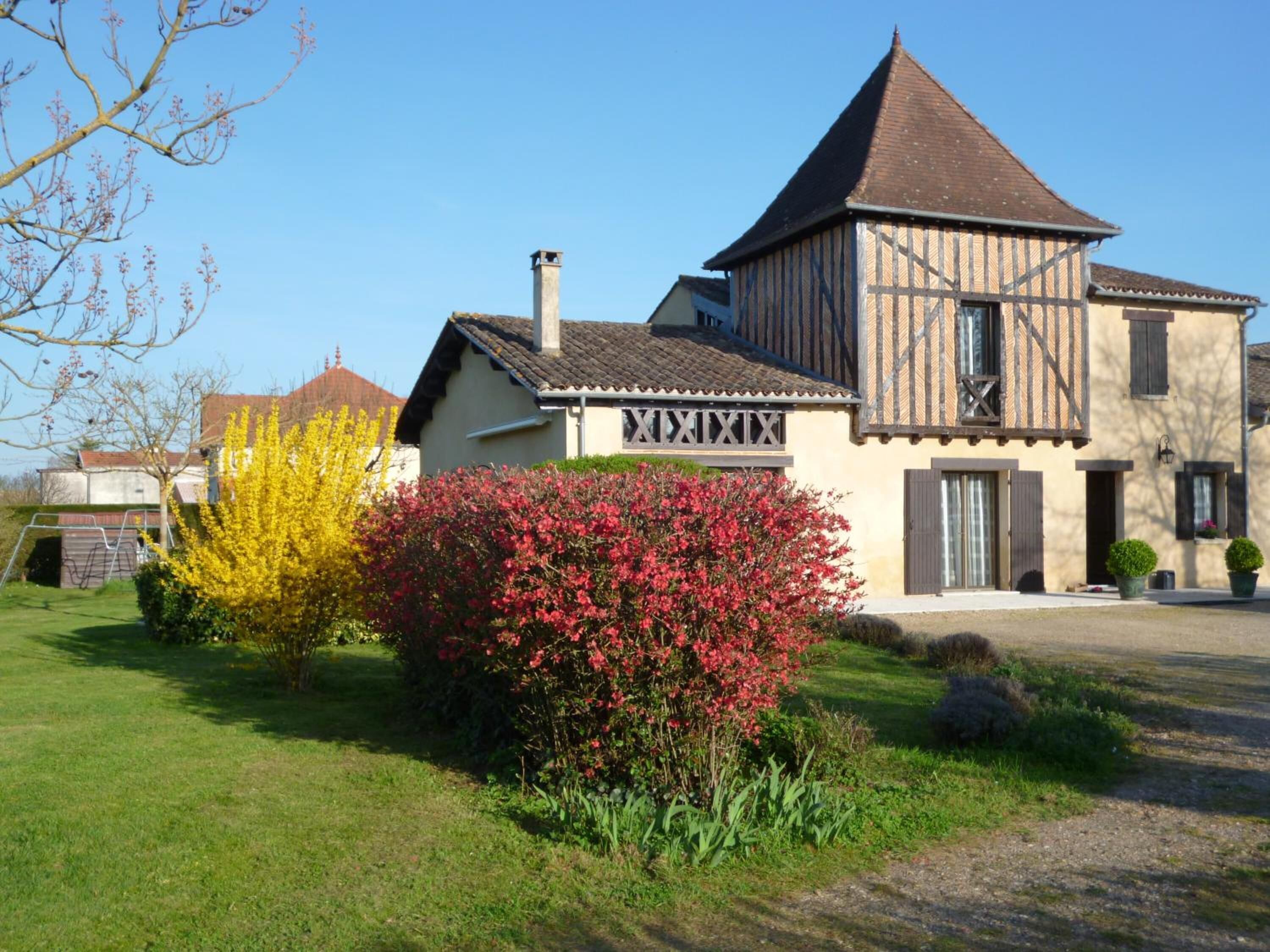A traditional half-timbered house with a prominent tower, surrounded by well-maintained gardens and a clear sky.