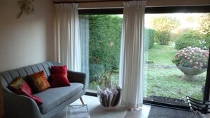 A living room with a grey sofa, red and yellow cushions, a glass coffee table, and a view of a garden through a glass door. - Les Sureaux (Le Pizou)