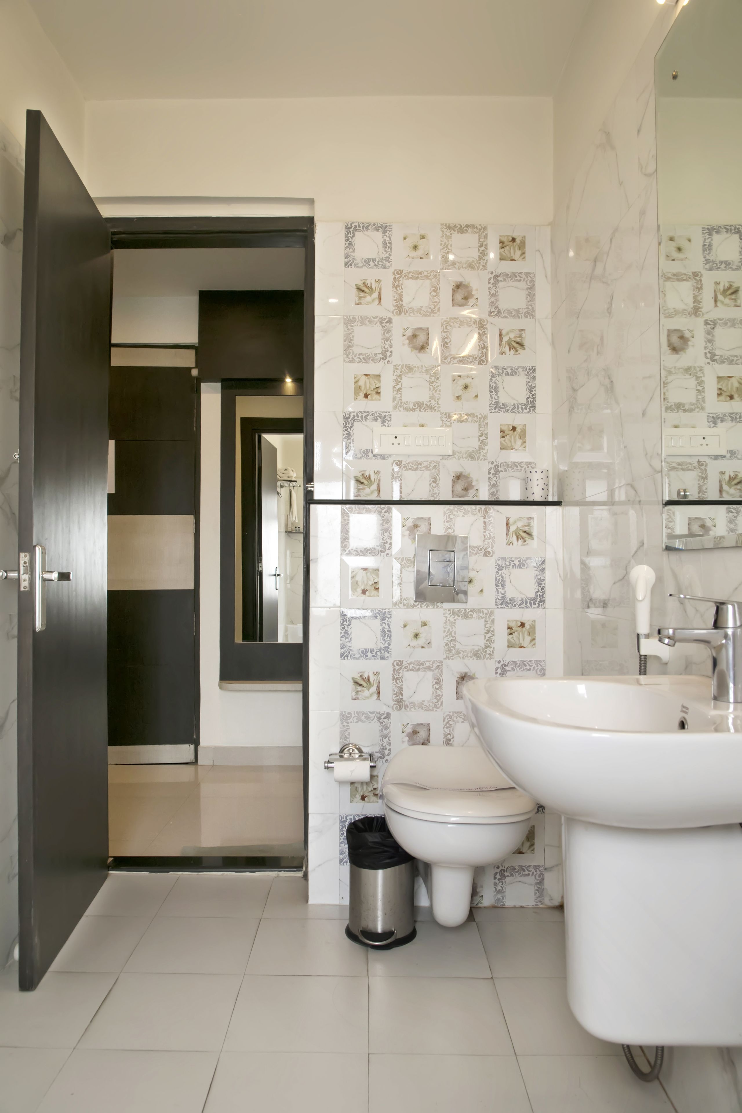 Un baño moderno con un espejo grande, lavamanos blanco y un inodoro. Las paredes están decoradas con azulejos estampados.