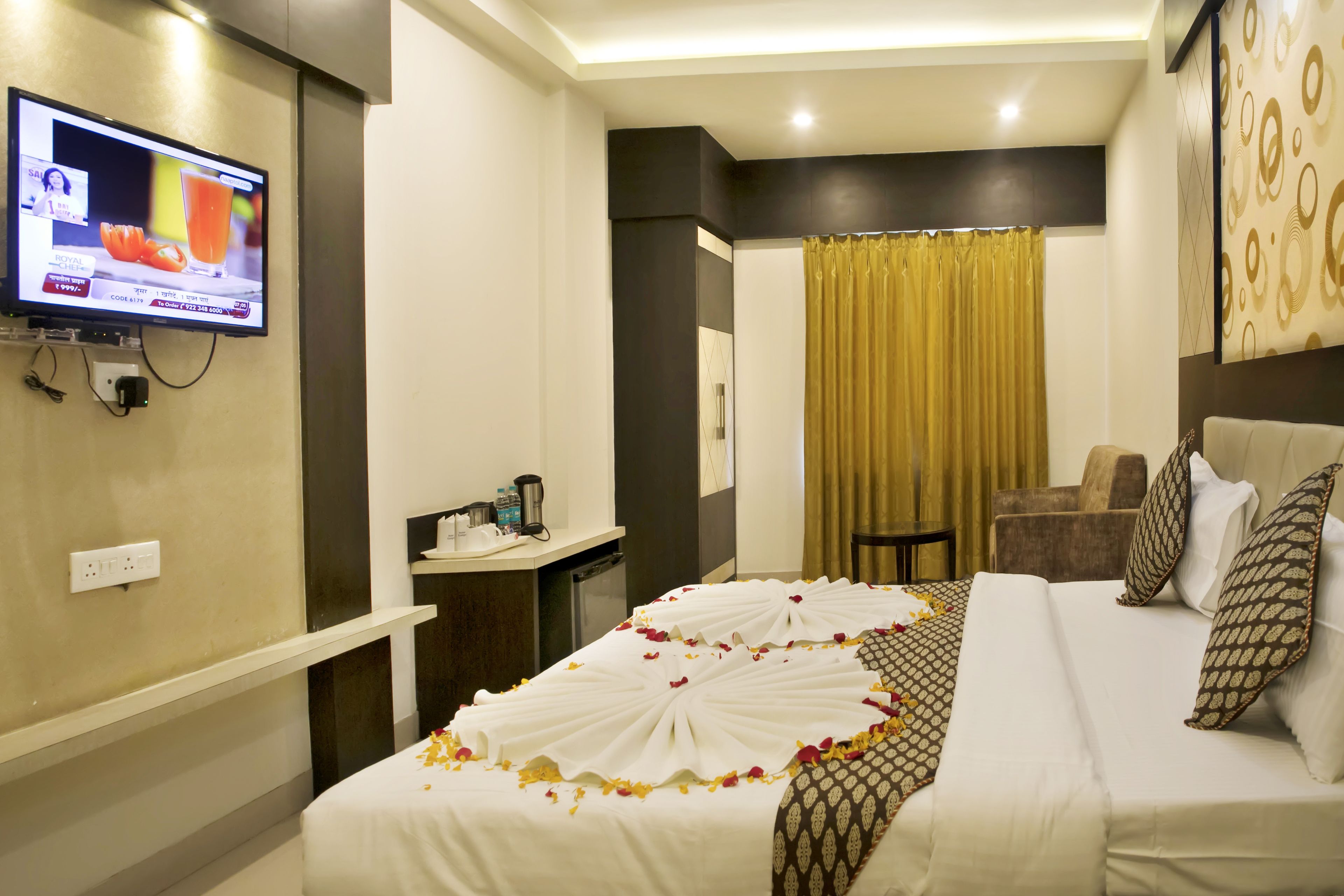 Habitación de hotel con una cama decorada con pétalos de rosa, un televisor montado en la pared, un escritorio con teléfono y cafetera, y vistas al exterior a través de una cortina.