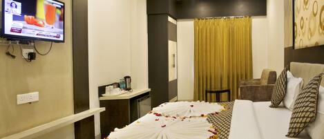 Habitación de hotel con una cama decorada con pétalos de rosa, un televisor montado en la pared, un escritorio con teléfono y cafetera, y vistas al exterior a través de una cortina.