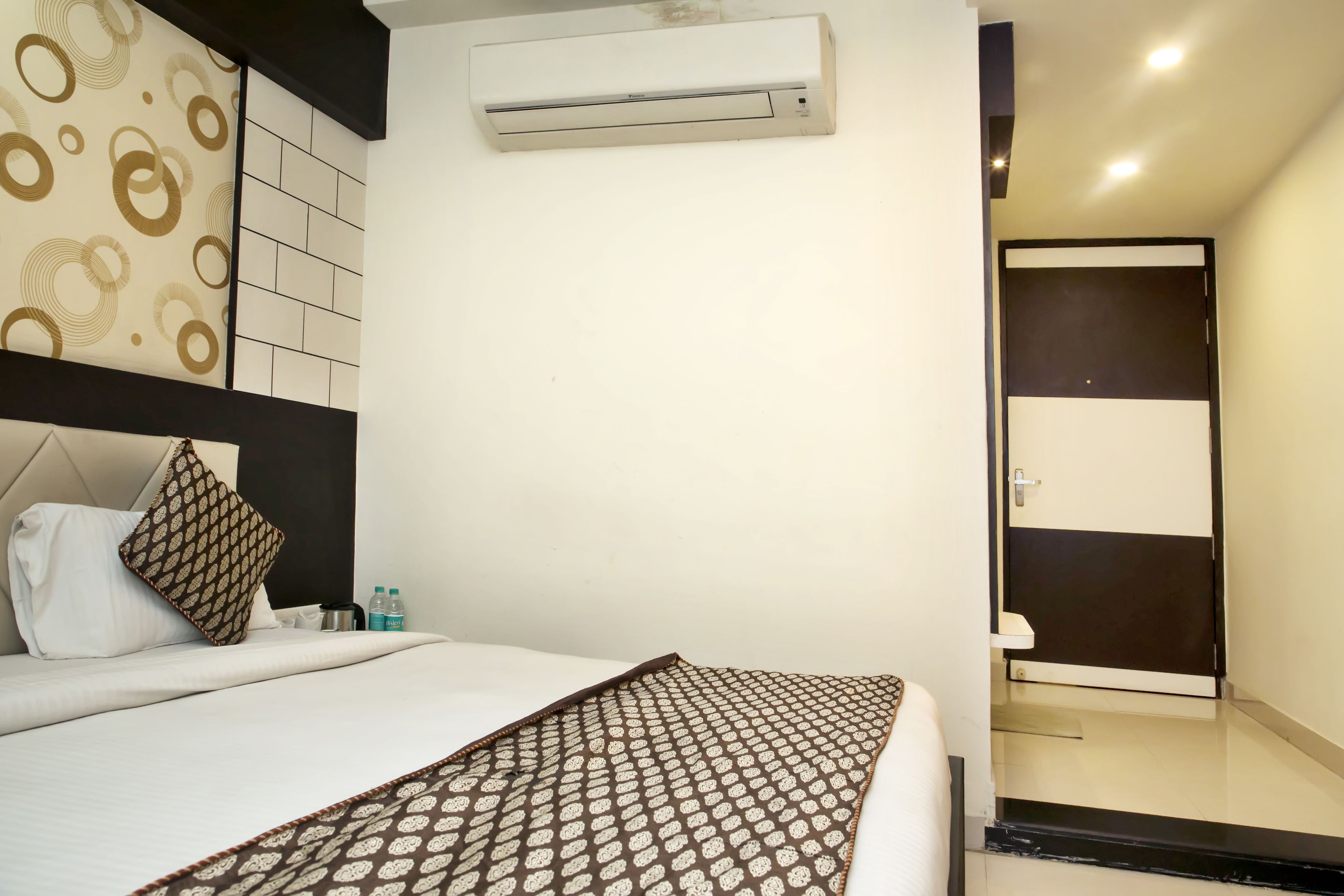 Habitación de hotel con una cama, un plaid estampado, un equipo de aire acondicionado, un baño con espejo y puerta, y una pared con diseño circular.