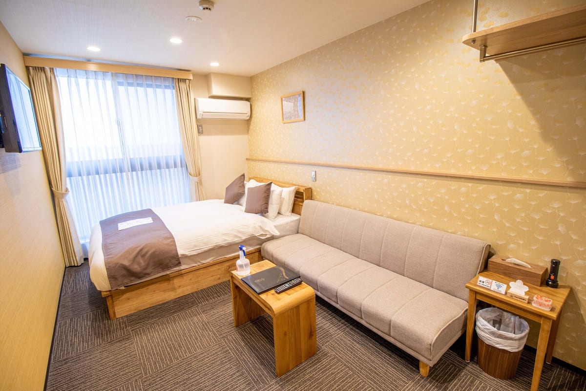 A hotel room with a bed, sofa, bedside table with a remote, a trash bin, and a small table with amenities.