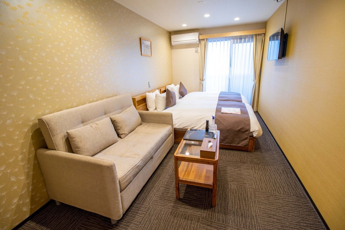 A hotel room with a sofa, two beds, a small wooden table, a TV, and an air conditioning unit.