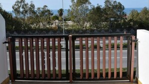 Uma varanda com portão de metal, com vista para uma rua arborizada e uma paisagem distante do mar.