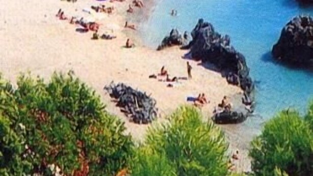 Uma praia com pessoas se bronzeando e nadando, cercada por rochas e vegetação.