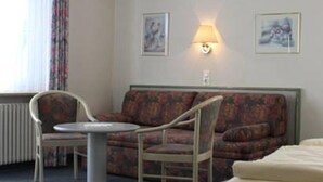 Hotellihuoneessa on sohva, kaksi nojatuolia, pieni pyöreä pöytä ja sänky. Seinällä on kaksi seinään kiinnitettyä lamppua ja kaksi kehystettyä kuvaa.