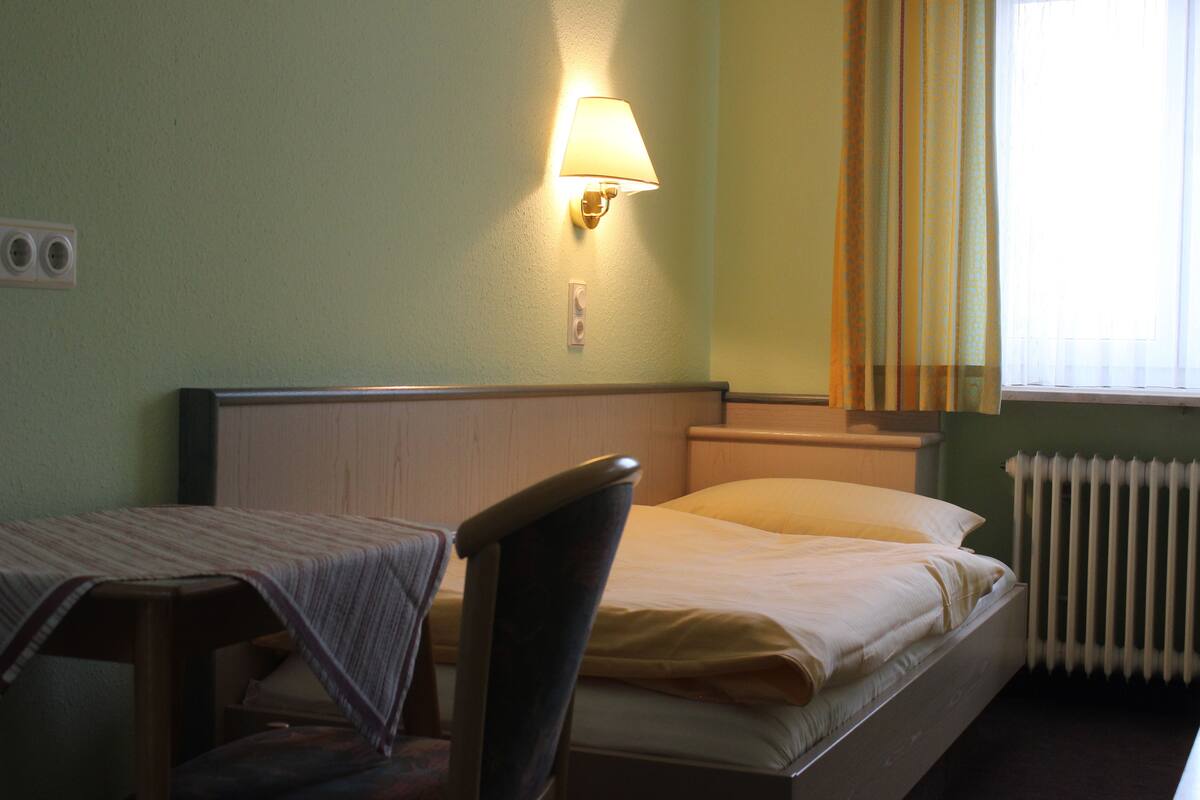 A single bed with a headboard, a bedside table with a lamp, and a window with curtains.