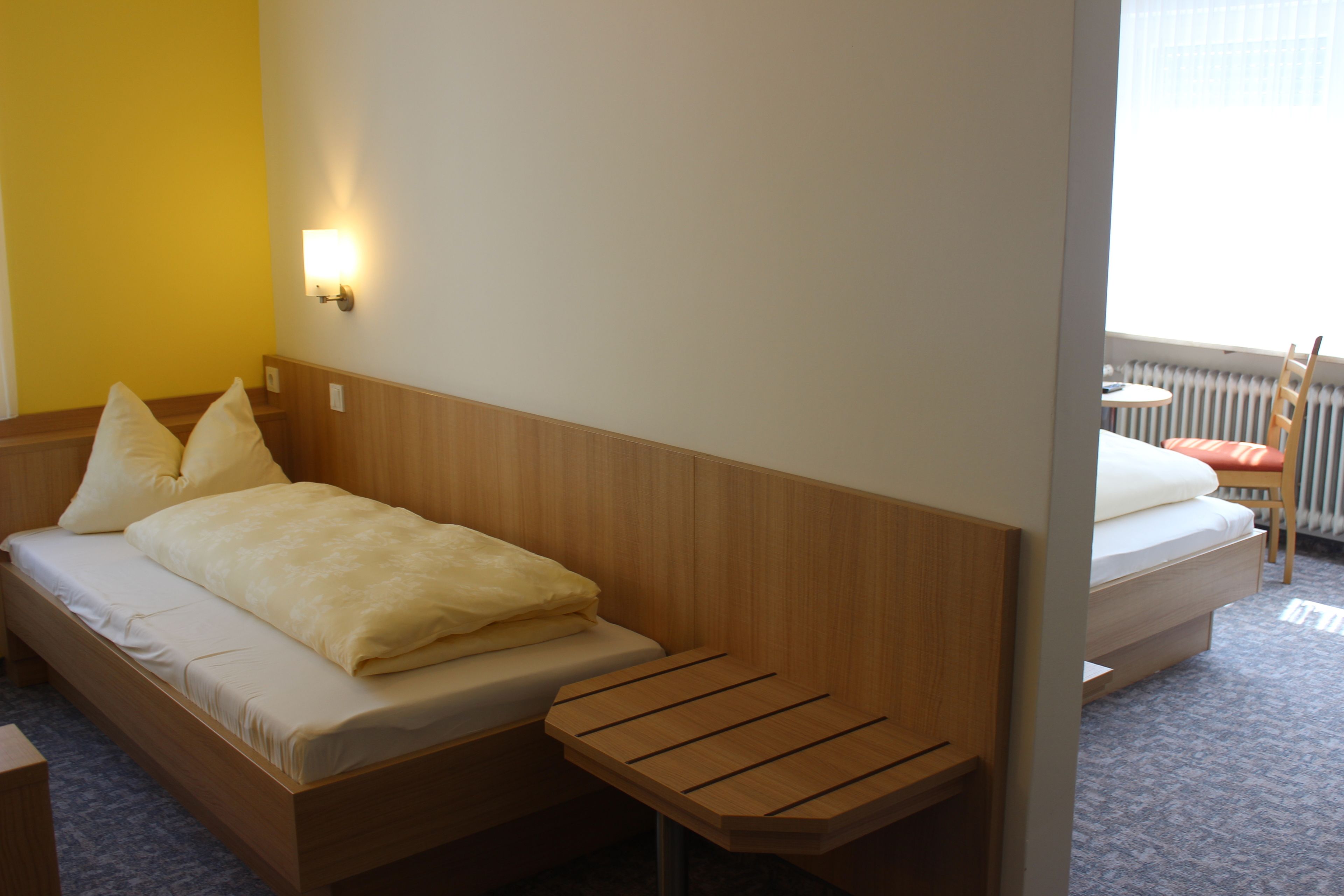 A hotel room with a wooden bed, a small wooden table, a wall-mounted light, and a view of the outdoors through a window.