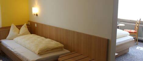 A hotel room with a wooden bed, a small wooden table, a wall-mounted light, and a view of the outdoors through a window.