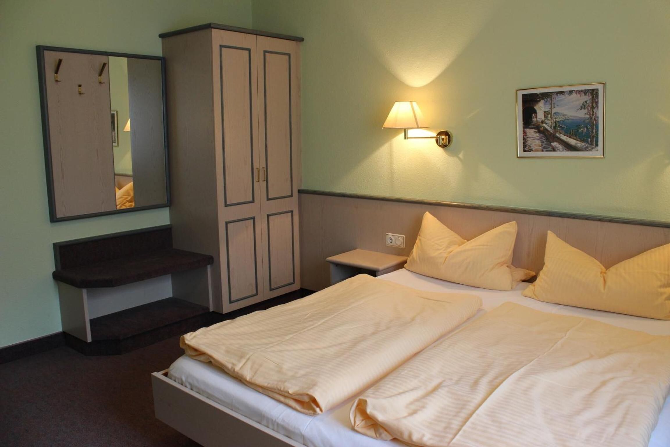 A hotel room with a bed, a nightstand, a mirror, a wardrobe, and a painting on the wall.
