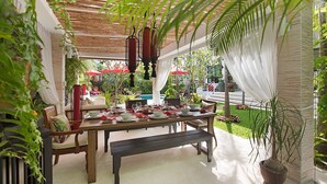 Uma área de refeições ao ar livre com uma mesa de madeira posta para quatro pessoas, rodeada por cadeiras. Há um banco no centro, e um vaso com planta visível à direita.