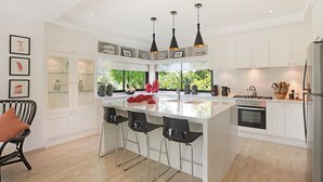 Cozinha moderna com armários brancos, uma ilha central com banquetas, iluminação pendente e vista para o verde lá fora.