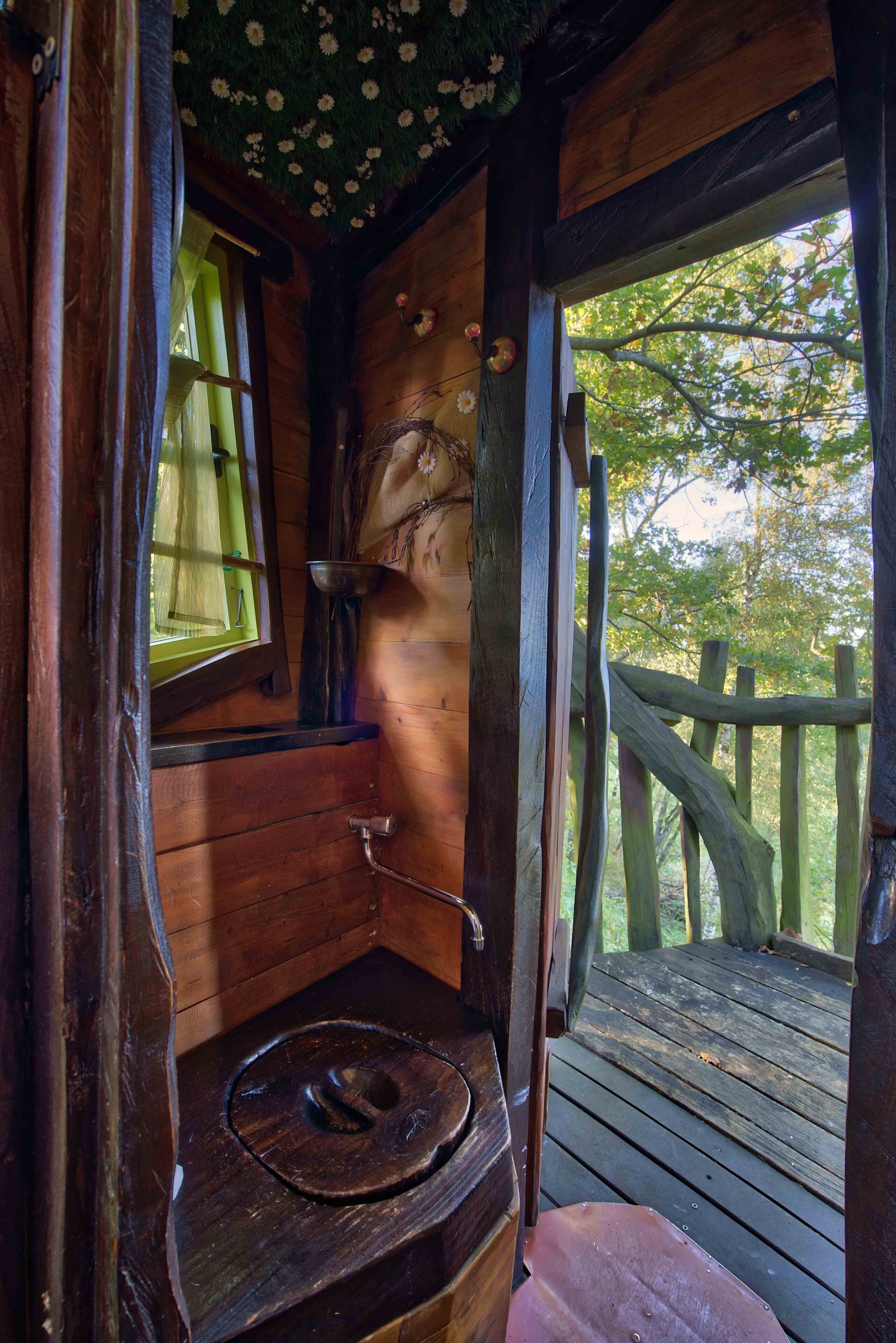 A rustic wooden cabin with a wooden toilet, a window with a green curtain, and a wooden deck.