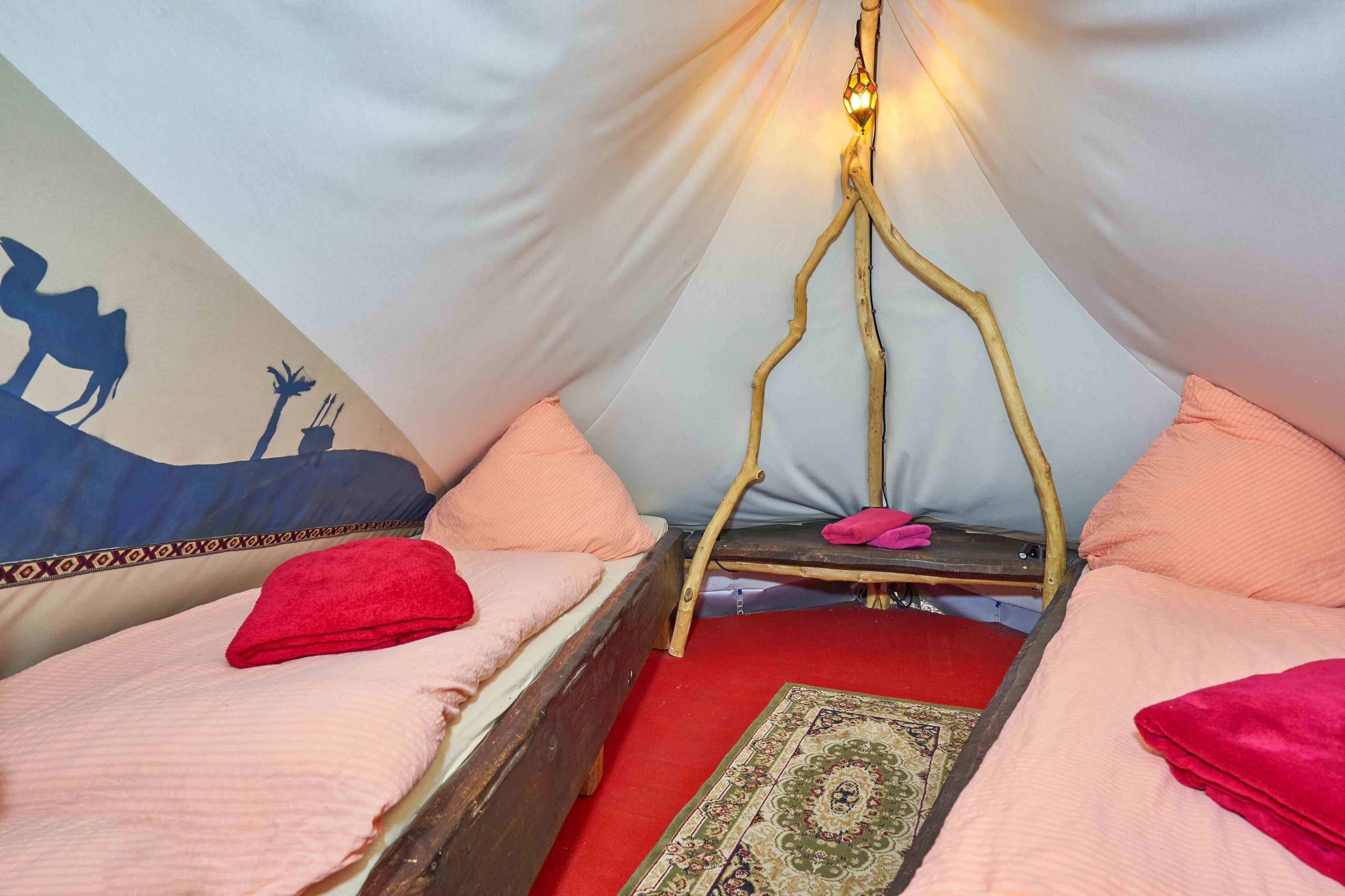 Um quarto em tenda com duas camas, cada uma com roupa de cama rosa e uma toalha vermelha. Uma pequena mesa com chinelos rosa fica entre as camas.