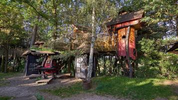 A rustic wooden structure with a thatched roof, a small wooden shed, and a treehouse with a red exterior.