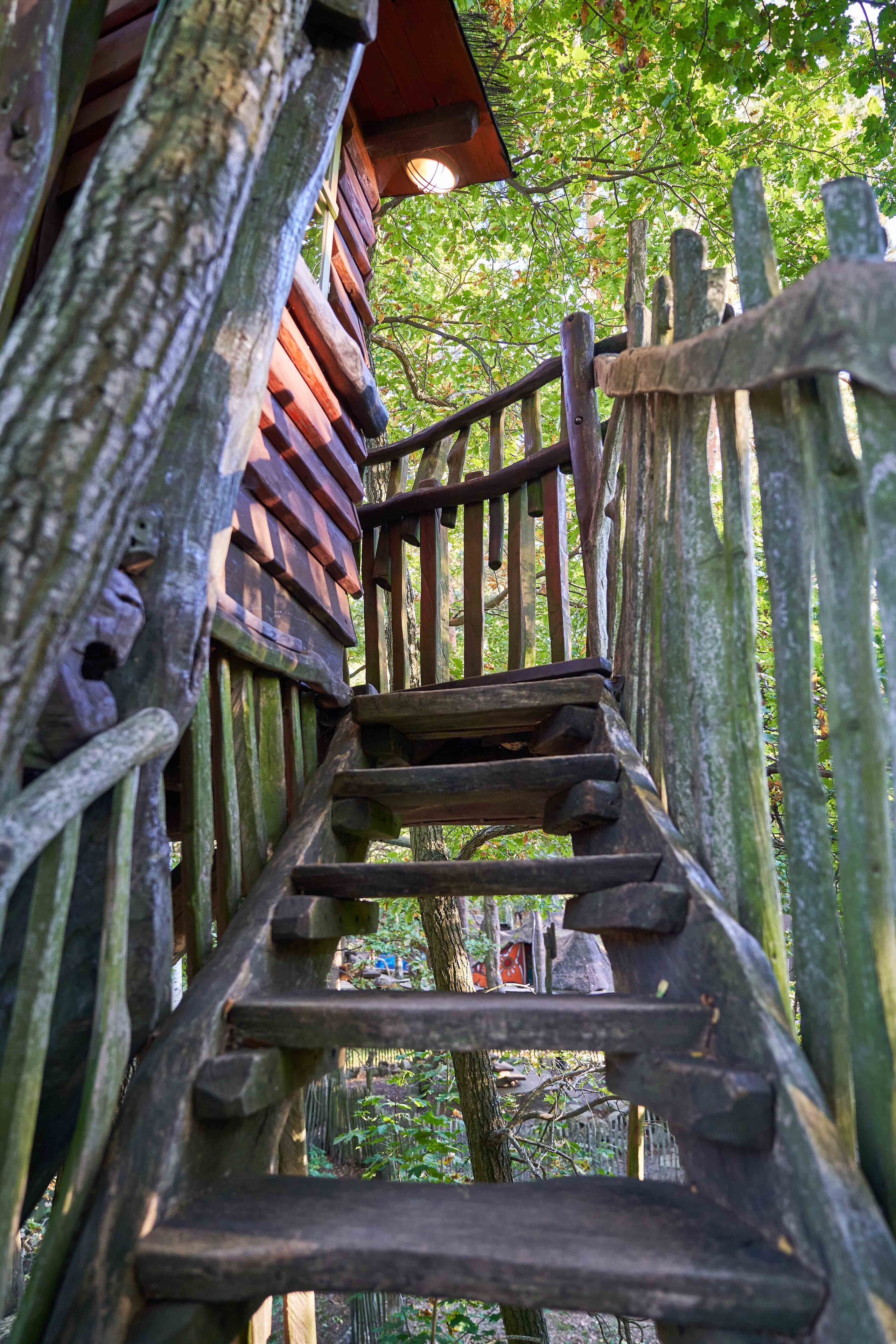 Una escalera de madera que sube hacia un edificio con techo rojo, rodeado de árboles.