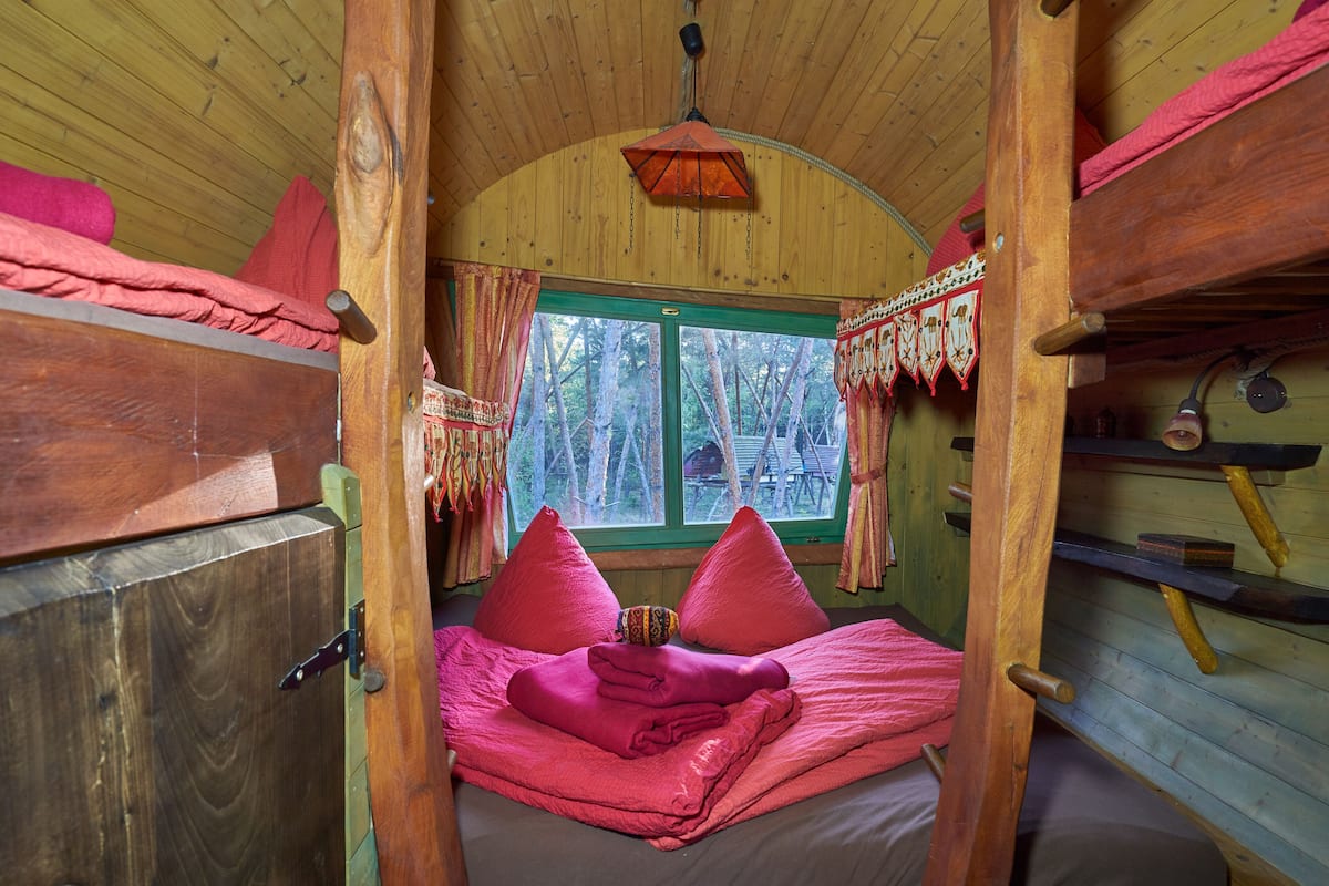 A cozy cabin bedroom with a wooden interior, a bed with pink bedding, a window with curtains, and a hanging light fixture.