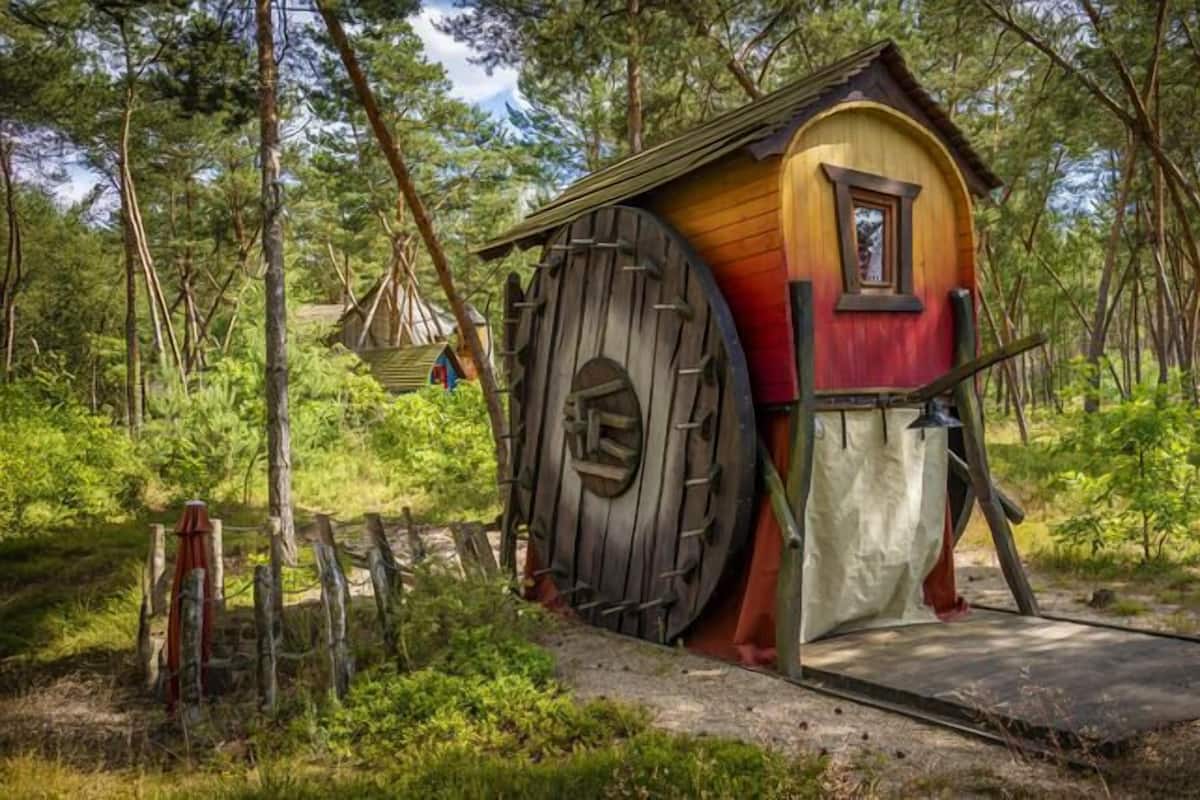 A wooden structure with a large wheel and a small window, surrounded by trees.