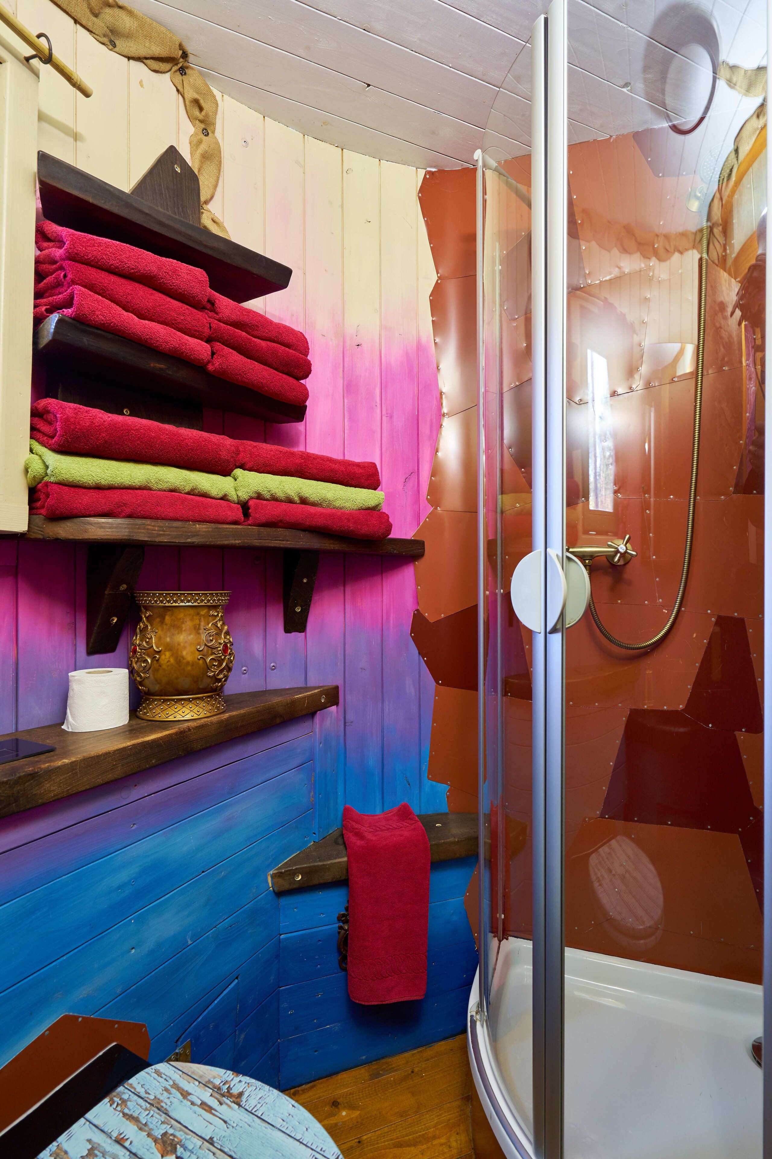 A bathroom with a glass shower enclosure, wooden shelves with towels, a toilet paper roll, and a decorative vase.