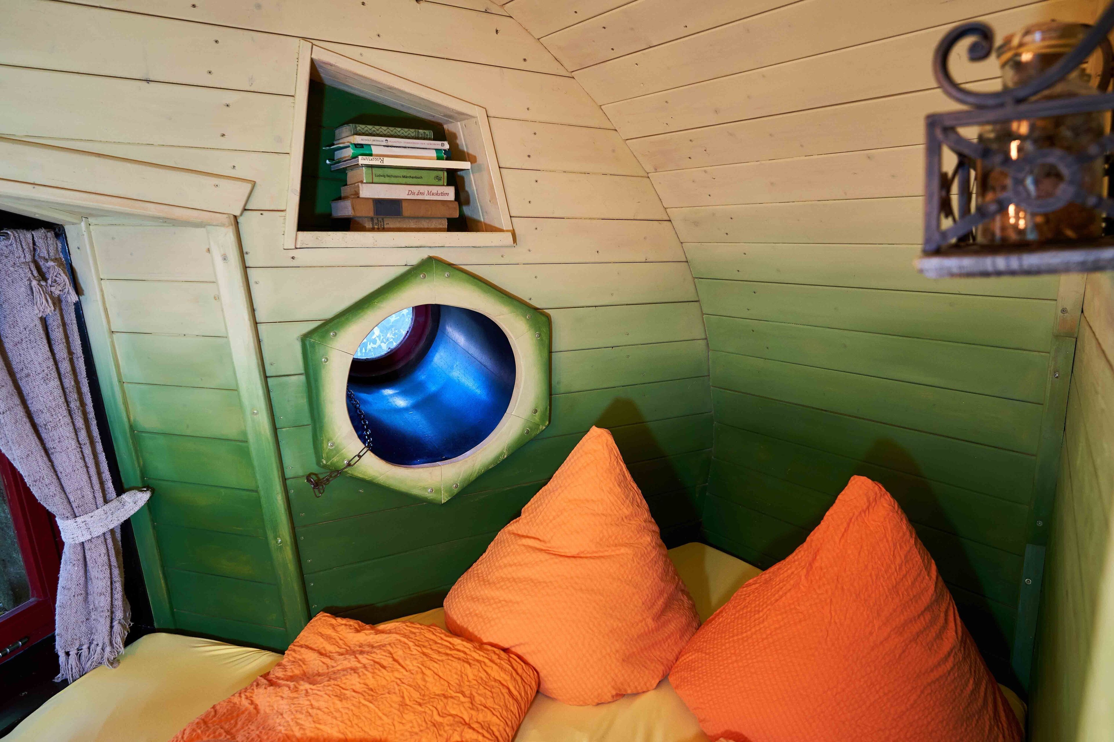A cozy room with a green and white color scheme, featuring a small window, a shelf with books, and orange pillows on the bed.
