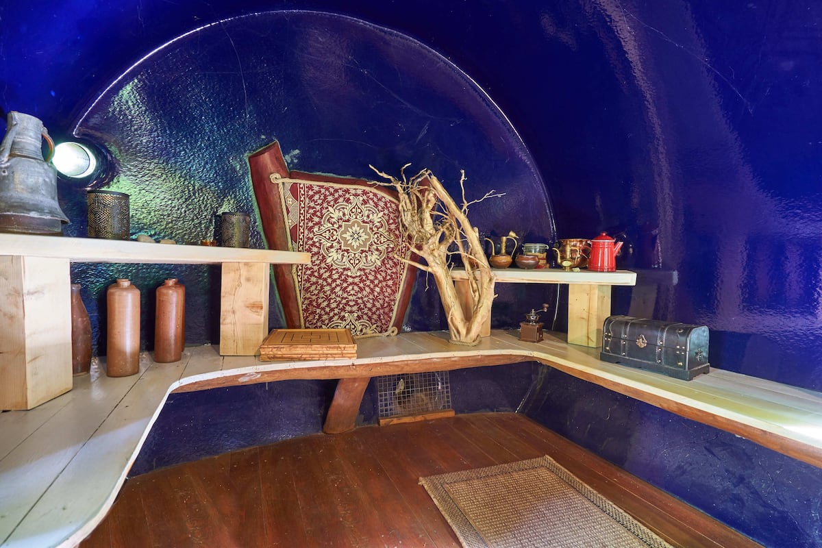 A room with a curved ceiling, a wooden table, a chair with a patterned cushion, and shelves with various items including a red teapot and small containers.