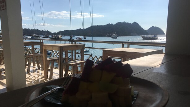 A dining area with a view of boats on water, wooden furniture, and a plate of fruit. - Bajo Sunset Hostel (Labuan Bajo)