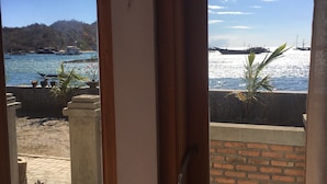 A view from a room with a wooden door and window, overlooking a beach with boats and a hilly landscape. - Bajo Sunset Hostel (Labuan Bajo)