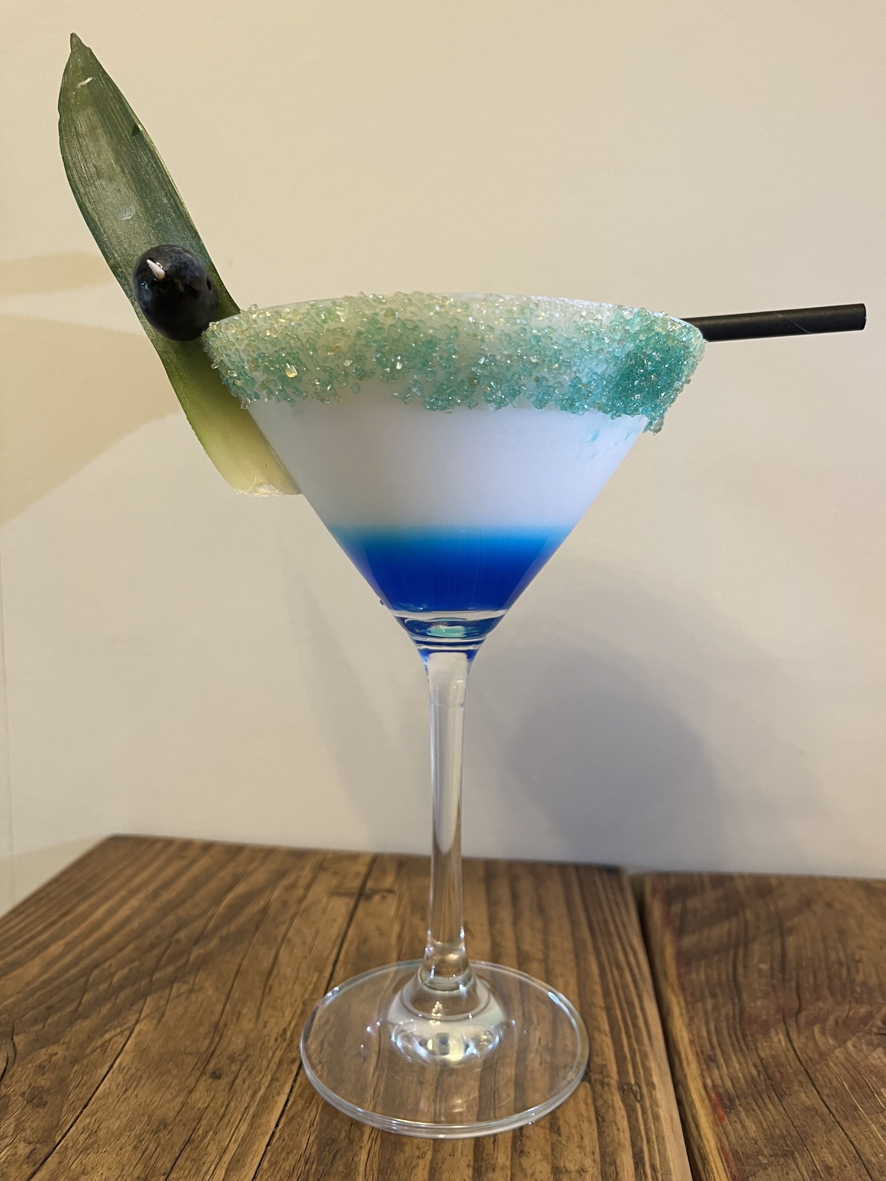 A martini glass with a blue and white layered drink, a sugar rim, and a green leaf garnish on a wooden table.