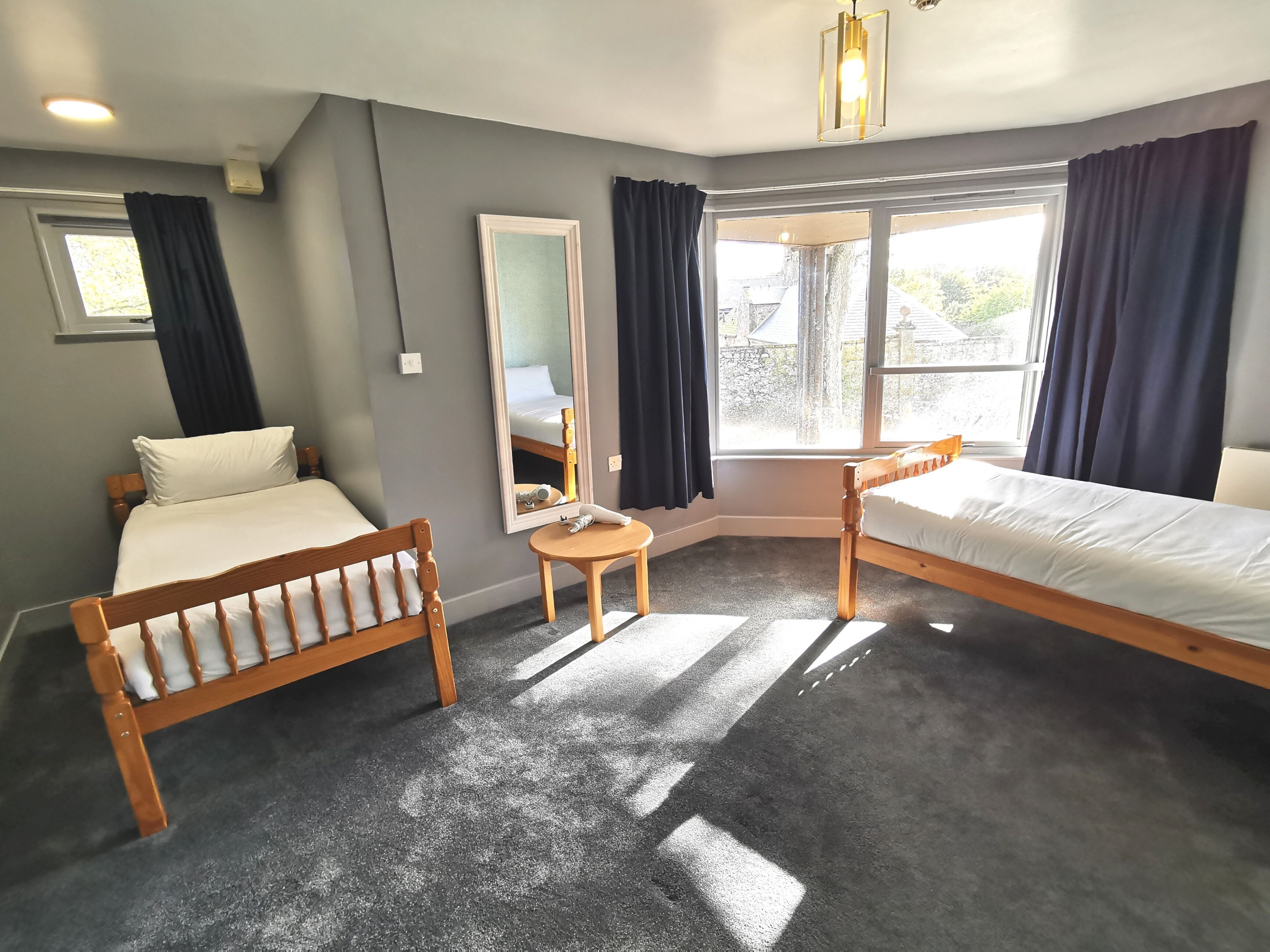 A hotel room with two wooden beds, a small round table, a mirror, and a window with curtains.