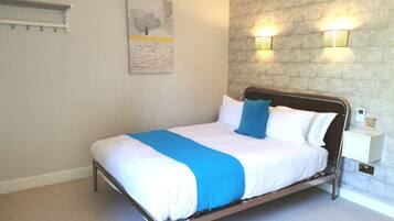 Una cama bien tendida con colcha azul y blanca, dos lámparas de pared y una mesita de noche con toma de corriente.