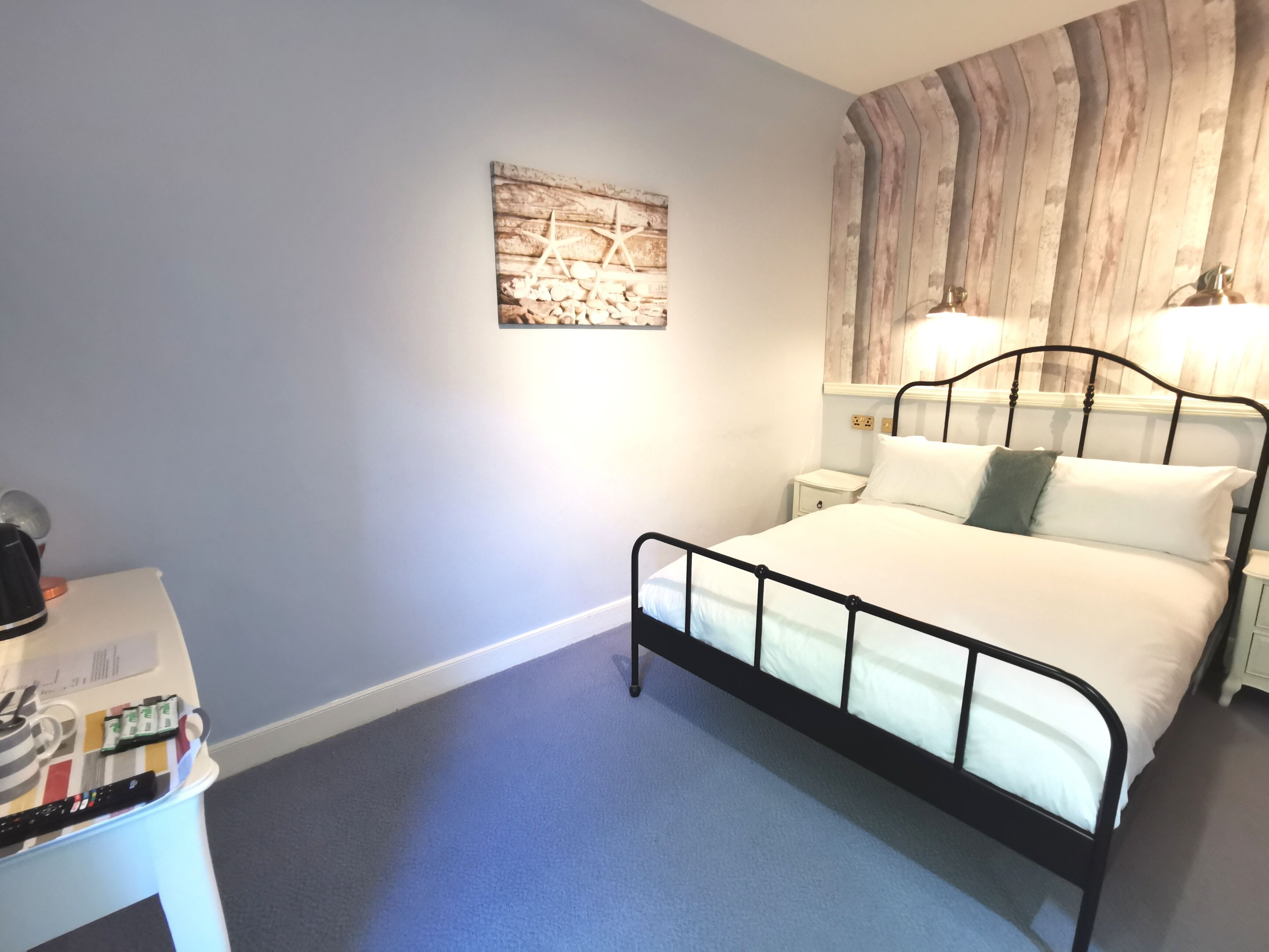 A bedroom with a bed, bedside tables, a nightstand, a wall-mounted lamp, and a small table with a kettle and a remote control.