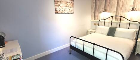 A bedroom with a bed, bedside tables, a nightstand, a wall-mounted lamp, and a small table with a kettle and a remote control.