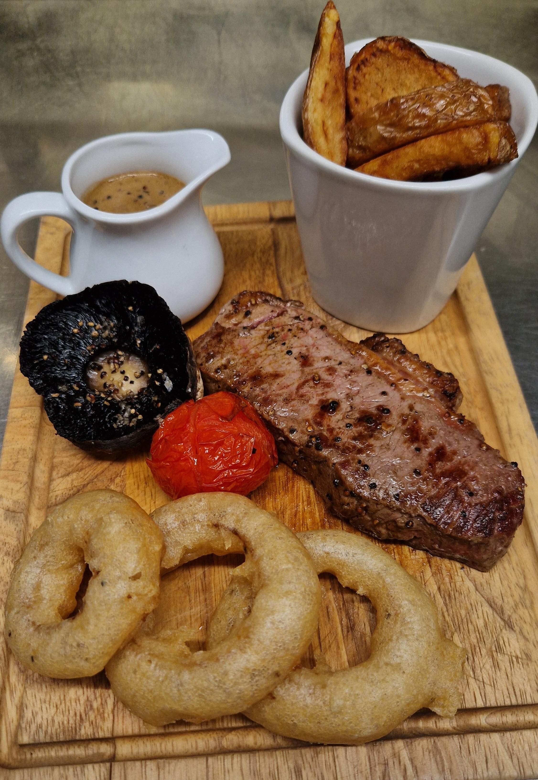A wooden cutting board with grilled steak, onion rings, grilled tomatoes, and a small white pitcher of sauce.