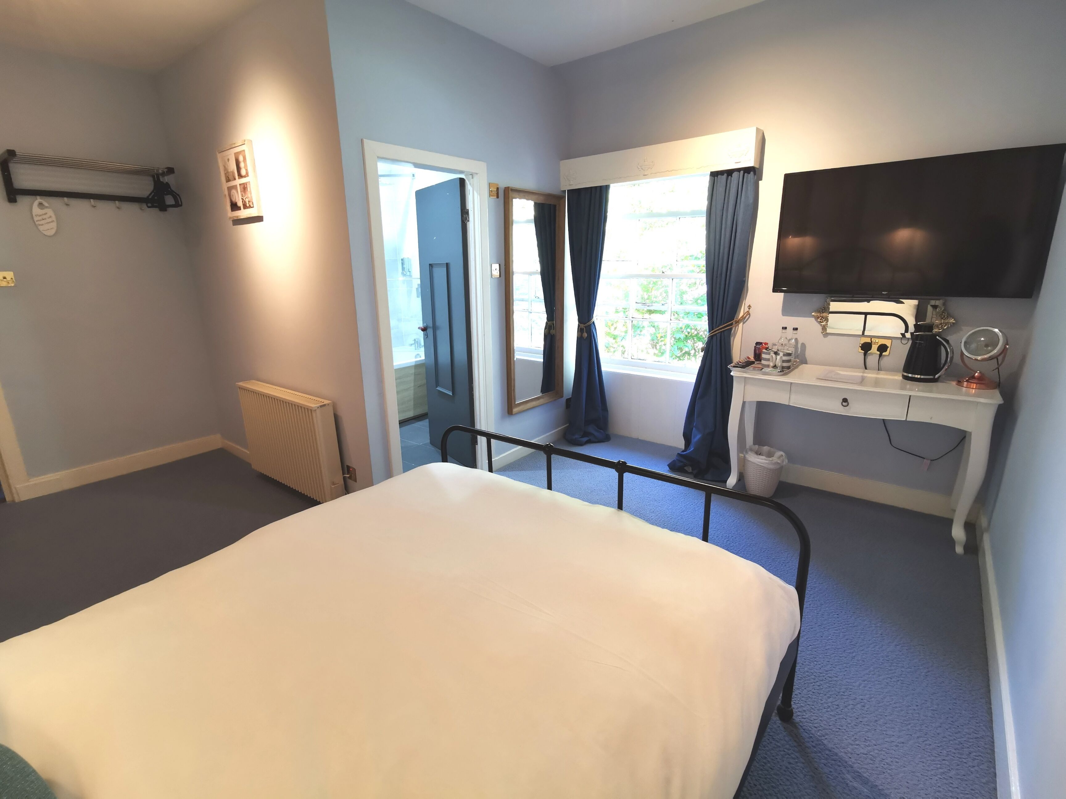Une chambre à coucher avec un lit, un bureau doté d’un miroir, un radiateur et une fenêtre avec des rideaux.